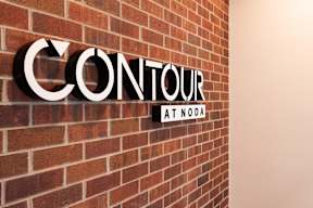 A sign that says "Contour at NODA" is mounted on a brick wall.