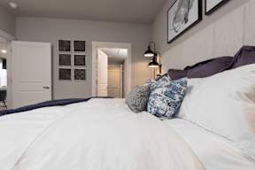 A bed with white sheets and a blue pillow is in the foreground of a room with a white door and a window.