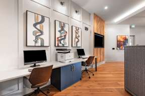A modern office with a wooden floor and white walls, featuring a desk with a computer and printer, a brown chair, and three abstract paintings on the wall.