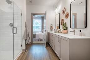 A bathroom with a walk-in shower, a large mirror, and a bedroom with a window.