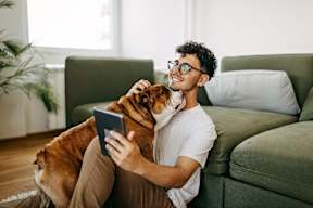 a man sitting on a couch holding a dog and looking at a tablet