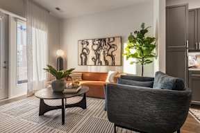 A living room with a grey armchair, a black coffee table, a potted plant, a sofa, and a painting on the wall.