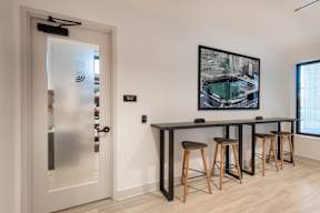 A modern bar area with a glass door, a framed picture on the wall, and four stools.