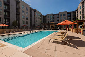 A swimming pool surrounded by lounge chairs and umbrellas in front of apartment buildings.