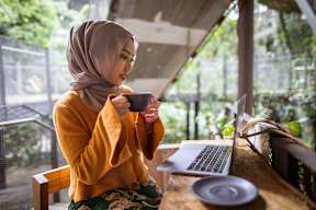 A woman in a yellow top and floral skirt is sitting at a wooden table with a laptop and a cup in front of her.