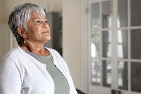 A woman with short grey hair wearing a white cardigan and a grey top stands in front of a white door.