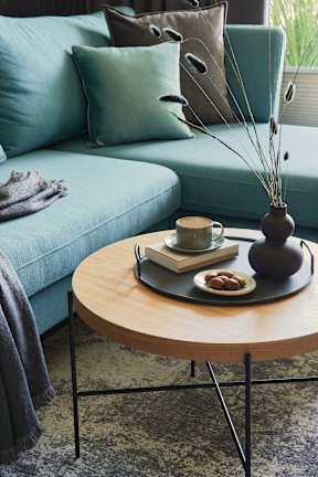 A living room with a blue couch, a wooden table with a vase, a book, a cup, and some fruit.