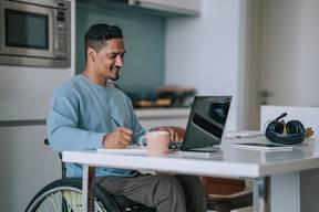 A man in a blue sweater is sitting at a table with a laptop and a cup of coffee.