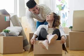 A man and woman are playing in a cardboard box in a room with other boxes and a couch.