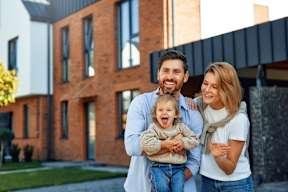 A family of three stands in front of a modern house.