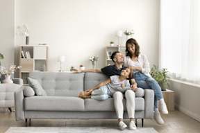 A woman and a child are sitting on a grey couch with a man lying on the floor.