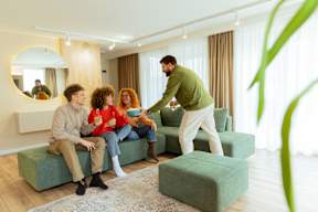 A man serving food to two people sitting on a green couch in a living room.