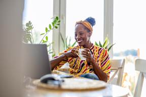 A woman in a colorful shirt is sitting at a table with a laptop and a cup in front of her.