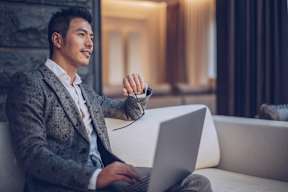 A man in a grey suit is sitting on a white couch using a laptop.