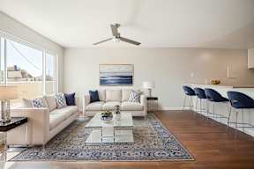 A modern living room with a white sofa set, a glass coffee table, and a ceiling fan.