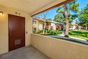 A brown door is on the left of a balcony with a view of a green lawn and trees.