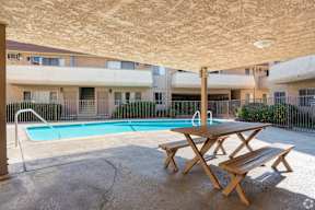 A wooden picnic table is in the middle of a concrete courtyard with a pool and apartment buildings in the background.