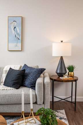 A living room with a grey couch, a side table with a lamp, and a painting of a bird on the wall.
