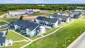 A large white building with a black roof is surrounded by smaller buildings and green grass.