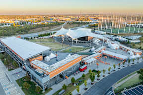 A large, modern building with a white and orange roof is surrounded by a parking lot and a grassy area.