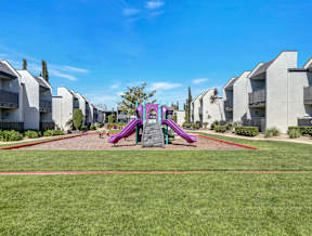 A playground with a purple slide in the middle of a grassy area with apartments in the background.
