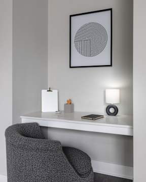 A grey chair is in front of a white desk with a lamp and a picture on the wall.