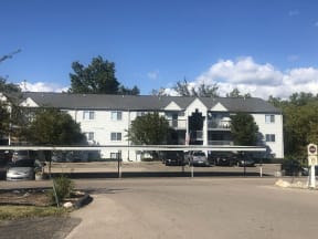 Drive into Greyberry Apartments with the building in the background.  The building is three stories and is grey in color with a dark grey roof. In between the drive and the apartment building is a grey carport. Behind the building are large green trees.