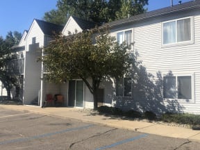 Two story grey apartment building with sidewalks leading up to the building.  There is a patio to the right of the breezeway. In front of the building is a tree and other green bushes.