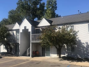Two story grey apartment building at Greyberry Apartments. There is a patio for the first-floor homes and a balcony on the second floor.  The breezeway has a staircase in it and is in the middle of the building.  There are two trees on either side of the breezeway and a sidewalk is in front of the building.