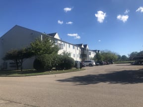Three story exterior of a grey apartment building.  There are balconies on the third floor of the building and lush green landscaping.  The building in to the left and to the right is a grey carport with cars parked under it.