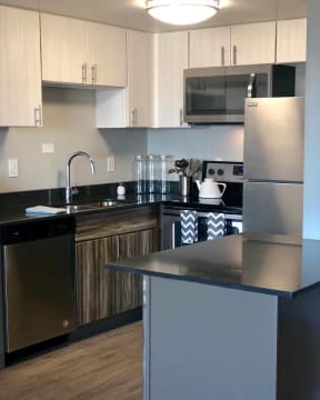 Image of a kitchen in the Jefferson. There are dark brown wood-like cabinets below and white cabinets on top. There are black countertops with stainless steel appliances. There is a sink to the left with a dishwasher and a stove and fridge on the right. The walls are a light grey with wood-like flooring.