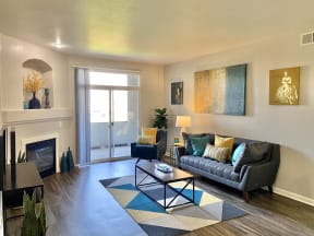 Windsor Denver Apartments - Interior View Of The Living Room Which Features Gold And Blue Artwork Hanging On The Wall, Large Glass Door That Leads Out To The Private Patio Space, Dark Navy Blue Sofa W