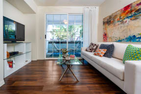 Living Room With Hardwood-Style Flooring, Sofa, Glass-Top Coffee Table, Media Cabinet with Glass Shelves, TV, And Sliding Glass Door Leading to Balcony. at Barrington Place, Rancho Cucamonga, CA