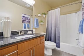 Apartments In Mountain View, CA - Americana Apartments - Bathroom With Large Vanity Mirror, Granite Countertop, Sink, Wood-Like Cabinets, Toilet, And A Shower With Tub
