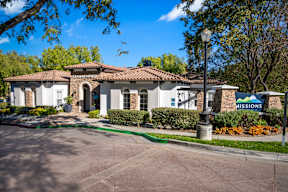 Pet-Friendly Apartments in Chino Hills, CA - Missions at Chino Hills - Leasing Center Exterior With Tile Roof, Arched Entry, Mission Sign, Landscaped Shrubs, And Tree Lined Driveway.