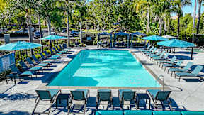 Three-Bedroom Apartments in Chino Hills, CA - Missions at Chino Hills - Resort Style Swimming Pool With Lounge Chairs, Teal Umbrellas, Shaded Cabanas, And Palm Trees In Sunny Courtyard.