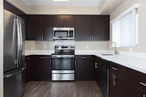 Palo Alto, California Apartments - Tan Plaza - Upgraded Kitchen with Stainless Steel Appliances, White Countertops, Dark Wood-Style Flat-Panel Cabinetry, Overhead Lighting, and Wood-Style Flooring.