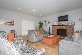 A living room with a grey couch, a grey armchair, a grey ottoman, a grey and white patterned rug, a wooden coffee table, a fireplace with a TV above it, and a potted plant.