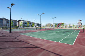 A tennis court with a red surface and white lines, surrounded by a fence and buildings in the background.