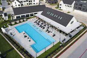 A large swimming pool with lounge chairs and umbrellas is surrounded by a white building and other buildings in the background.