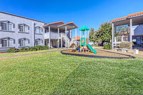 A playground with a green slide and a blue umbrella is in front of a building.