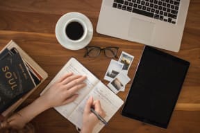 person writing in journal with laptop, notebook and cup of cofee