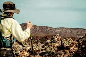 A man in a yellow jacket and hat is hiking on a rocky trail.