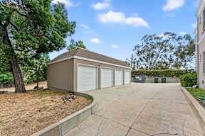 A detached garage with a brown roof and white doors is surrounded by a driveway and trees.