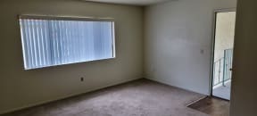 Carpeted living room with large window and vertical blinds at Magnolia Apartments in Riverside, Califonia.