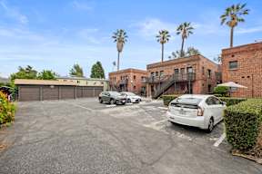 A white car is parked in a parking lot with a building and palm trees in the background.