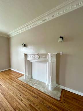 A white fireplace with a wooden mantle and a white marble hearth sits in a room with wood floors and white walls.