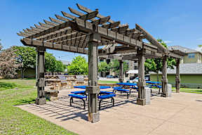 A wooden pergola with blue benches is situated in a park.