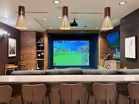 A room with a long table and chairs in front of a large screen displaying a golf game.