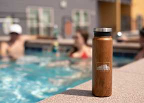 A brown water bottle with a silver lid sits on the edge of a pool.